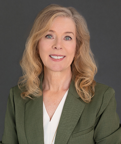 Becky Ringenberg, with shoulder-length blonde hair, wears a green blazer over a white blouse as she poses confidently in front of a plain dark background.