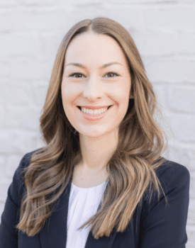 Tana Massey, with her long brown hair cascading gracefully, smiles warmly in a black blazer and white blouse against a light-colored brick wall.