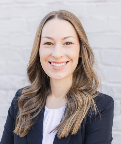 Tana Massey, with her long brown hair cascading gracefully, smiles warmly in a black blazer and white blouse against a light-colored brick wall.