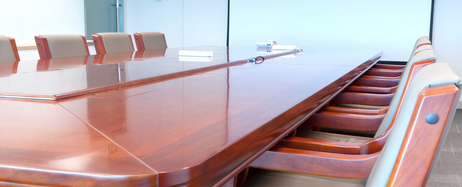 A long, polished wooden conference table with empty chairs is arranged in a meeting room, ready to strengthen your board of directors with strategic meetings against the backdrop of a blank screen.