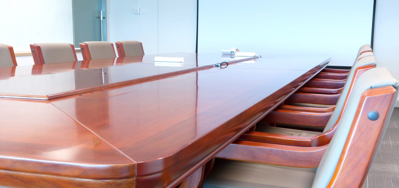 A long, polished wooden conference table with empty chairs is arranged in a meeting room, ready to strengthen your board of directors with strategic meetings against the backdrop of a blank screen.