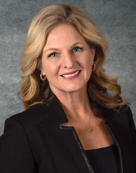 Jeanne Bernick, with her wavy blonde hair, smiles warmly while wearing a black blazer and a gold necklace against a textured gray background.