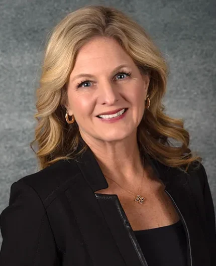 Jeanne Bernick, with her wavy blonde hair, smiles warmly while wearing a black blazer and a gold necklace against a textured gray background.