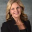 Jeanne Bernick, with her wavy blonde hair, smiles warmly while wearing a black blazer and a gold necklace against a textured gray background.