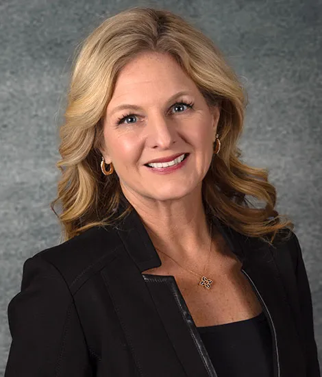 Jeanne Bernick, with her wavy blonde hair, smiles warmly while wearing a black blazer and a gold necklace against a textured gray background.