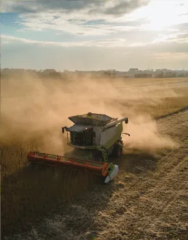 A combine harvester works in a field under a cloudy sky, creating dust clouds as it harvests crops.