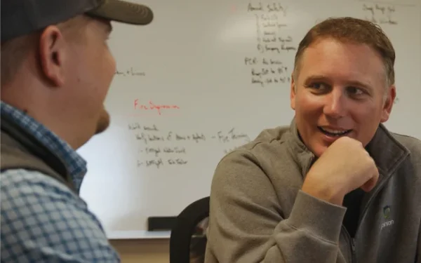 Two men sit and converse in an office. One is wearing a cap, the other has his hand on his chin. A whiteboard with notes is visible in the background.