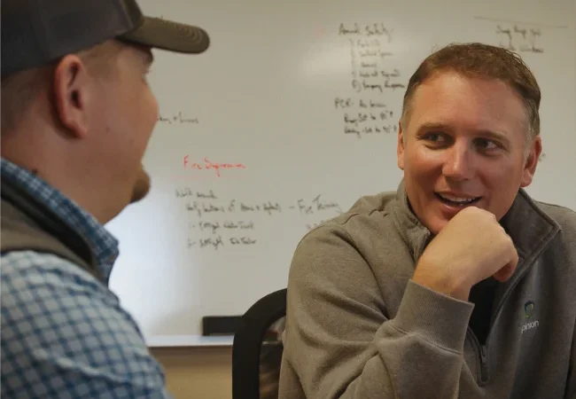 Two men sit and converse in an office. One is wearing a cap, the other has his hand on his chin. A whiteboard with notes is visible in the background.