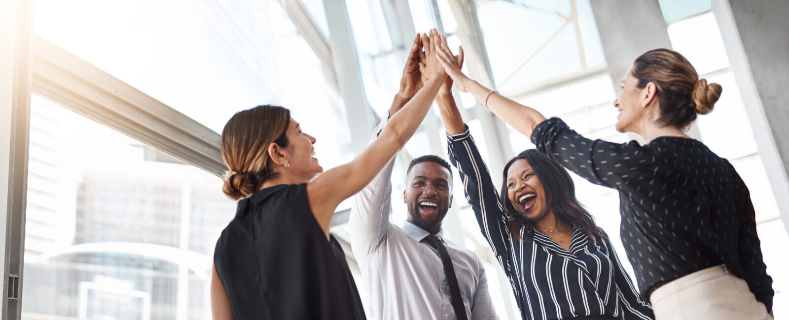 Four colleagues in business attire high-fiving in a bright office setting, smiling and celebrating.