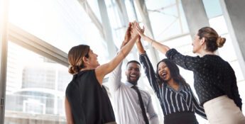 Four colleagues in business attire high-fiving in a bright office setting, smiling and celebrating.