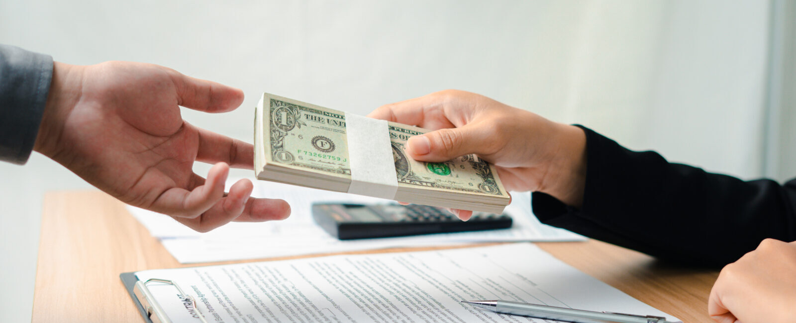 One person hands a stack of U.S. dollar bills to another across a table with documents, a pen, and a calculator.