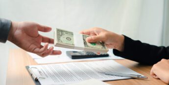 One person hands a stack of U.S. dollar bills to another across a table with documents, a pen, and a calculator.