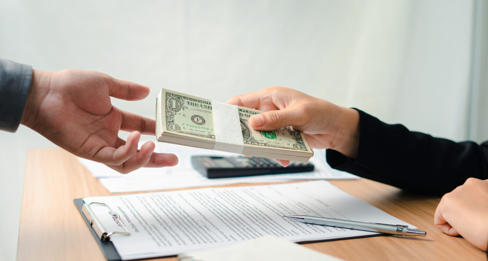 One person hands a stack of U.S. dollar bills to another across a table with documents, a pen, and a calculator.