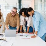 A group of six people is smiling and looking at a laptop in a modern office, where papers and notebooks are scattered on the table—perhaps exploring 6 strategies to cultivate a positive work environment.