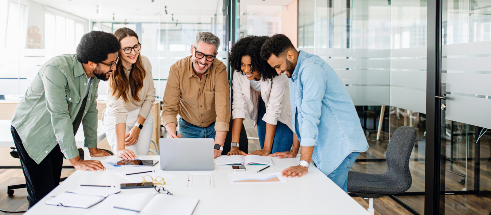 A group of six people is smiling and looking at a laptop in a modern office, where papers and notebooks are scattered on the table—perhaps exploring 6 strategies to cultivate a positive work environment.