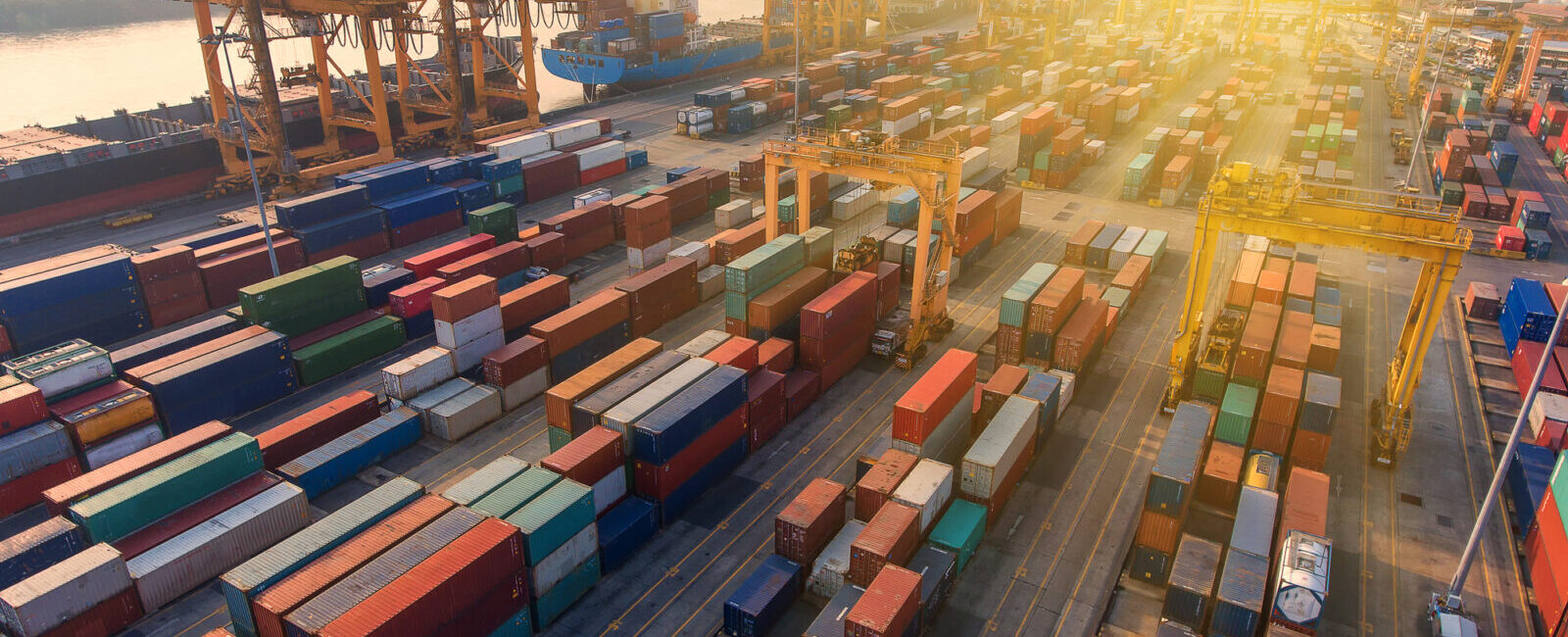 Aerial view of a busy shipping port at sunset with numerous colorful shipping containers and large cranes along a dock near the water.
