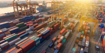 Aerial view of a busy shipping port at sunset with numerous colorful shipping containers and large cranes along a dock near the water.