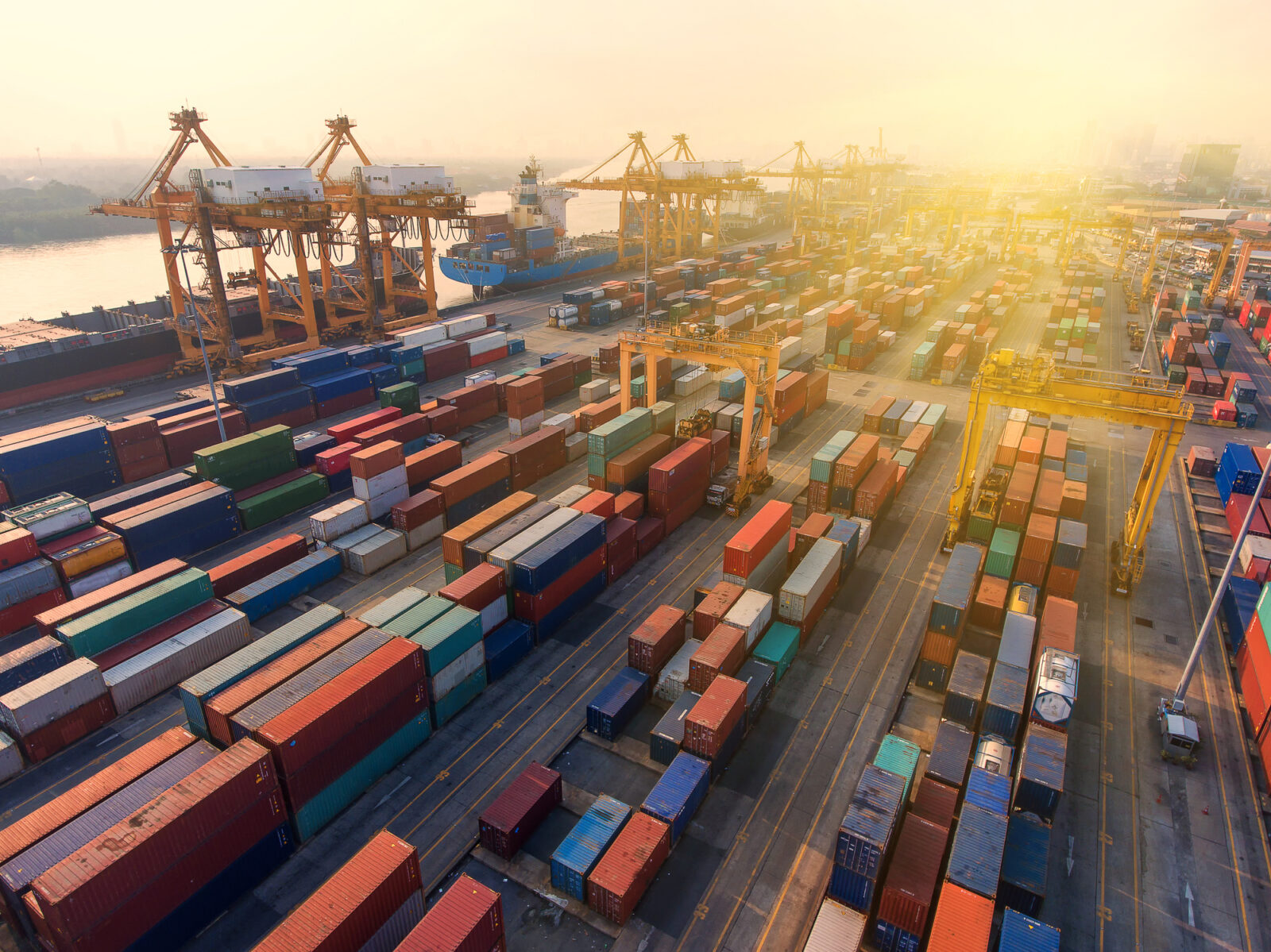 Aerial view of a busy shipping port at sunset with numerous colorful shipping containers and large cranes along a dock near the water.