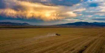 A lone vehicle drives across a vast, flat field under a dramatic, cloudy sky at sunset, with mountains in the background.