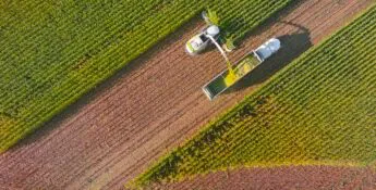 Aerial view of a combine harvester offloading crops into a truck on a dirt road, surrounded by green fields.