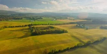Aerial view of a vast rural landscape with fields, scattered trees, distant hills, and a partially cloudy sky.