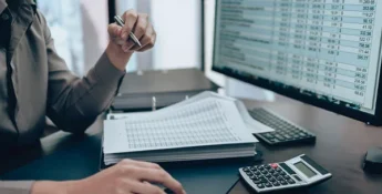 Person working at a desk with a computer displaying spreadsheets, holding a pen. Papers, keyboard, and calculator are also on the desk.