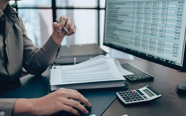 Person working at a desk with a computer displaying spreadsheets, holding a pen. Papers, keyboard, and calculator are also on the desk.