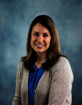 Amy with long brown hair, wearing a blue blouse and beige cardigan, poses against a blue-gray background, exuding confidence as if ready for her next transactional advisory meeting.