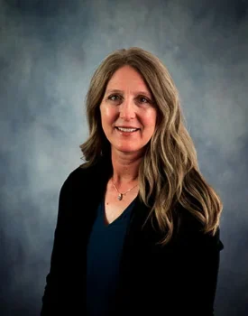 Erin with long hair and a blue shirt, wearing a black jacket, poses against a gray backdrop, exuding a confident air akin to someone in transactional advisory.