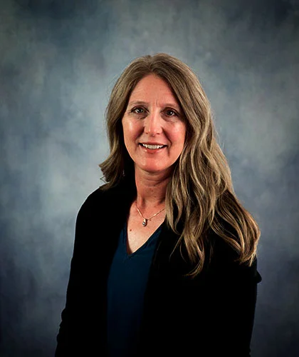 Erin with long hair and a blue shirt, wearing a black jacket, poses against a gray backdrop, exuding a confident air akin to someone in transactional advisory.