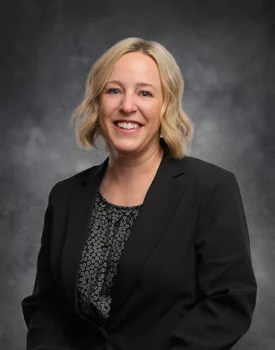 Brea Bauer, with short blonde hair, smiles warmly as she dons a black blazer over a patterned blouse. She stands elegantly against a dark, neutral backdrop.