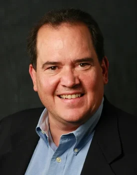A smiling Chris in a suit jacket and blue shirt stands confidently against a dark background, embodying the professionalism of transactional advisory.