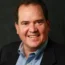 A smiling Chris in a suit jacket and blue shirt stands confidently against a dark background, embodying the professionalism of transactional advisory.