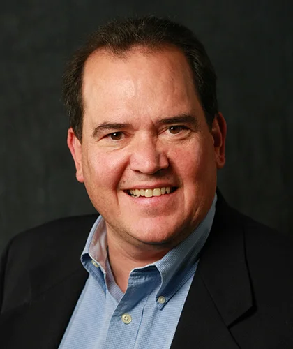 A smiling Chris in a suit jacket and blue shirt stands confidently against a dark background, embodying the professionalism of transactional advisory.