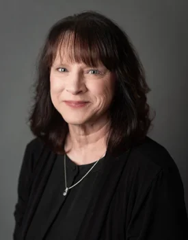 Brenda White, A woman with shoulder-length dark hair, donning a black sweater and necklace, smiles confidently against a gray background, embodying the essence of transactional advisory excellence.