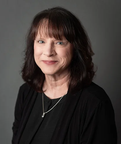 Brenda White, A woman with shoulder-length dark hair, donning a black sweater and necklace, smiles confidently against a gray background, embodying the essence of transactional advisory excellence.