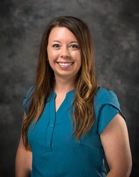 Dana with long brown hair smiles at the camera, exuding confidence in a teal short-sleeve blouse against a dark gray textured background, embodying the essence of transactional advisory expertise.
