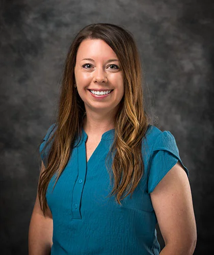 Dana with long brown hair smiles at the camera, exuding confidence in a teal short-sleeve blouse against a dark gray textured background, embodying the essence of transactional advisory expertise.