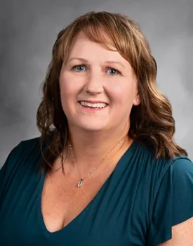 A smiling Shellee with shoulder-length wavy hair and a teal top stands poised against a grey background, exuding the confidence of someone experienced in transactional advisory.