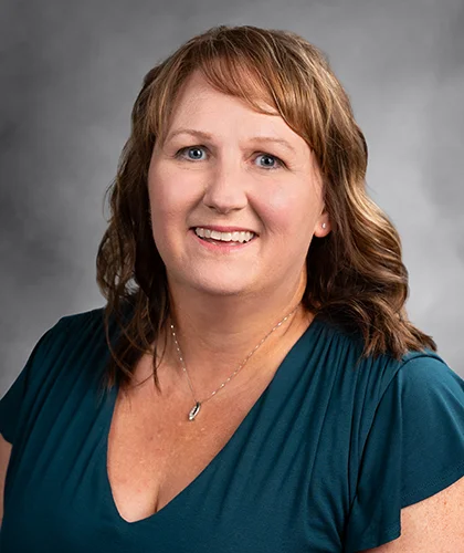 A smiling Shellee with shoulder-length wavy hair and a teal top stands poised against a grey background, exuding the confidence of someone experienced in transactional advisory.