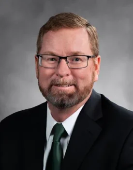Steve with glasses, a beard, and short hair, dressed in a suit and tie, exudes confidence in his professional headshot against a grey background. His welcoming smile reflects the expertise he brings to transactional advisory services.