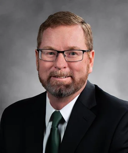 Steve with glasses, a beard, and short hair, dressed in a suit and tie, exudes confidence in his professional headshot against a grey background. His welcoming smile reflects the expertise he brings to transactional advisory services.