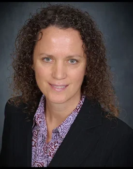 Christie Bettencourt, with her curly hair, dons a black blazer and patterned shirt, offering a subtle smile against a dark backdrop.
