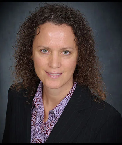 Christie Bettencourt, with her curly hair, dons a black blazer and patterned shirt, offering a subtle smile against a dark backdrop.