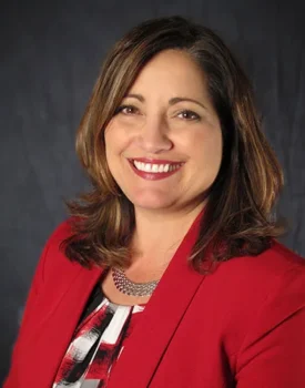 Geri smiling, wearing a red blazer and patterned top, exuding confidence against a dark backdrop, embodies the essence of transactional advisory expertise.