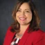 Geri smiling, wearing a red blazer and patterned top, exuding confidence against a dark backdrop, embodies the essence of transactional advisory expertise.