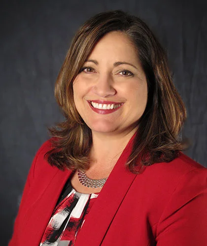 Geri smiling, wearing a red blazer and patterned top, exuding confidence against a dark backdrop, embodies the essence of transactional advisory expertise.