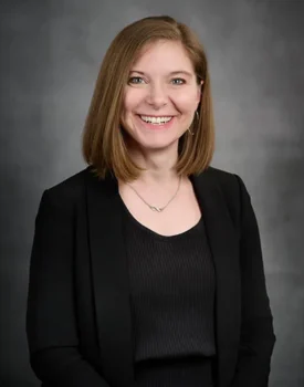 Jill with shoulder-length brown hair, wearing a black blazer and top, smiles confidently against a gray background, exuding the poised professionalism often associated with transactional advisory.