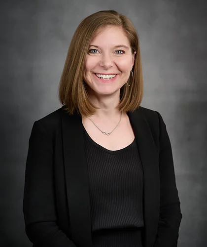 Jill with shoulder-length brown hair, wearing a black blazer and top, smiles confidently against a gray background, exuding the poised professionalism often associated with transactional advisory.