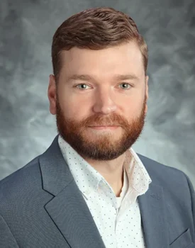 Kevin with short brown hair and a beard is wearing a gray suit jacket and a white polka-dot shirt, embodying the confidence of transactional advisory expertise, posing against a gray textured background.